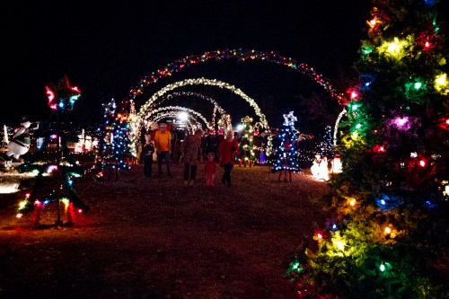 A festive night scene with colorful holiday lights, archways, and families walking through a decorated park.