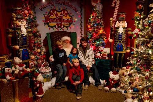 Four children pose together in a festive Christmas setting with decorations, nutcracker figures, and a Santa backdrop.