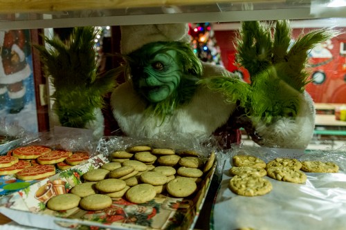 The Grinch peeks through a glass display filled with various holiday cookies.