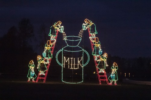 Colorful holiday lights depict children pouring milk from ladders into a large milk container against a dark sky.