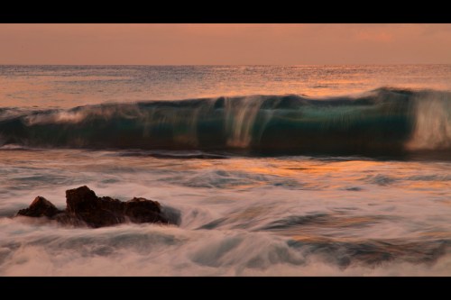 A serene ocean wave crashes against a rocky shore at sunset, with soft colors reflecting on the water's surface.