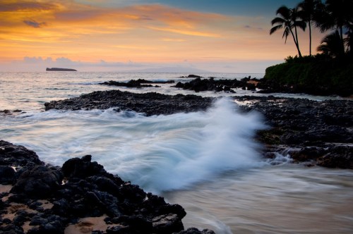 Sunset over a rocky beach with gentle waves and palm trees silhouetted against a colorful sky.