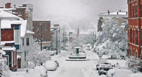 A snowy street scene with a fountain, surrounded by snow-covered buildings and parked cars. Visibility is low due to snowfall.