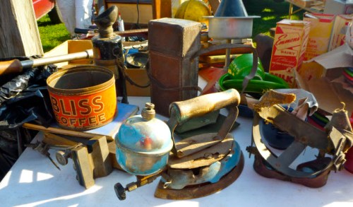 A collection of vintage kitchen tools and containers, including coffee cans, irons, and a kettle, displayed on a table.