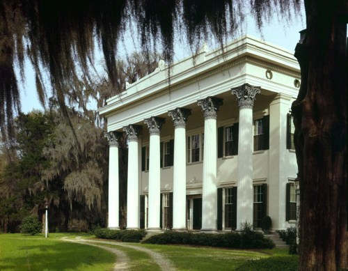 A grand, white-columned mansion surrounded by greenery and trees, featuring classic architectural details.