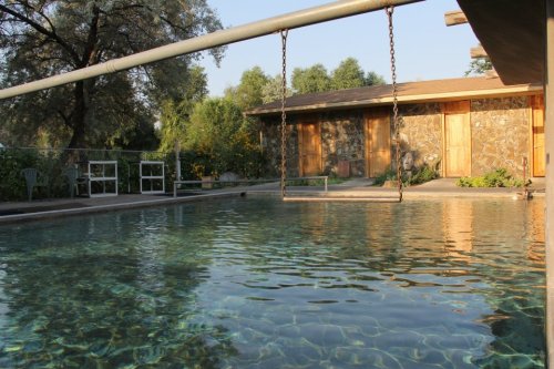 A serene pool with clear water, a swing nearby, and a stone building surrounded by greenery in the background.