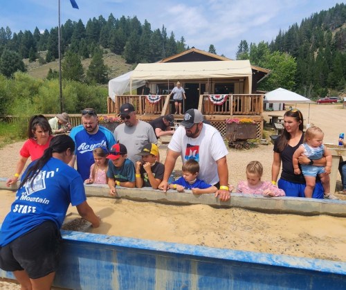 Families and children enjoy a gem panning activity at an outdoor event, with a rustic building in the background.