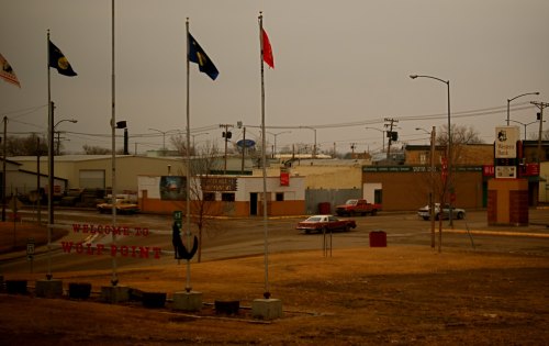 A view of a small town with flags, a welcome sign, and a vintage car parked on a quiet street.