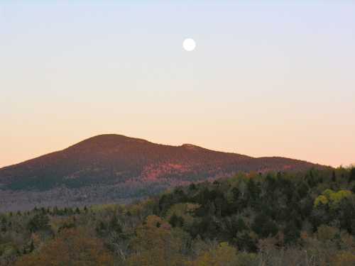 A serene mountain landscape at dusk, with a full moon rising above the peaks and colorful autumn foliage below.