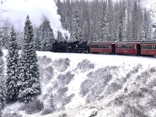 A steam train with red carriages travels through a snowy landscape, surrounded by evergreen trees.
