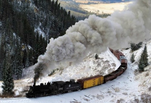A steam locomotive chugs along snowy tracks, billowing smoke amidst a forested landscape and rolling hills.