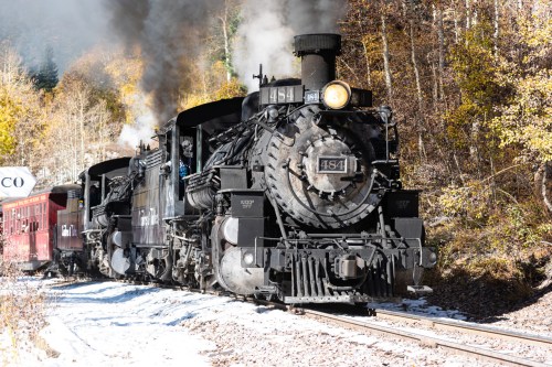 A vintage steam locomotive chugs along a scenic track surrounded by autumn foliage. Smoke billows from its chimney.