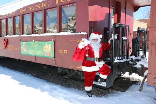 Santa Claus poses next to a snow-covered train car, wearing a red suit and waving cheerfully.