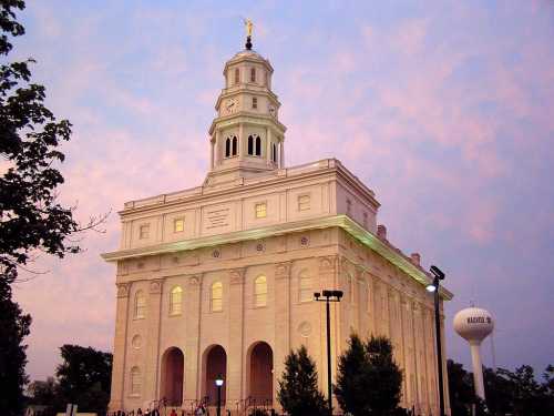 Historic building with a clock tower, illuminated against a twilight sky, surrounded by trees and a water tower nearby.