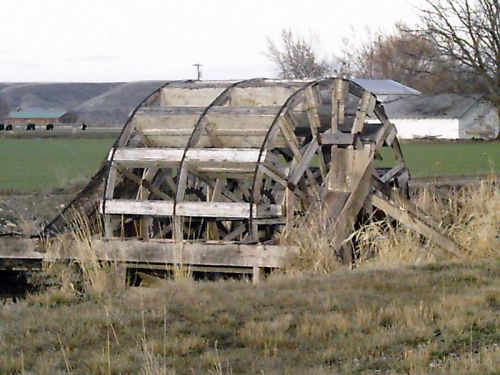 A weathered wooden waterwheel sits in a grassy field, surrounded by sparse vegetation and distant buildings.