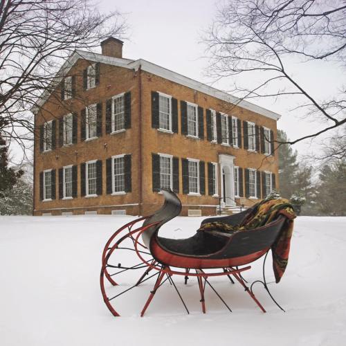 A historic brick building with black shutters, surrounded by snow, features a red sleigh with a colorful blanket in the foreground.