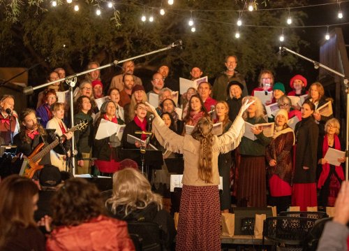 A large choir performs outdoors under string lights, with a conductor leading and audience members seated in front.