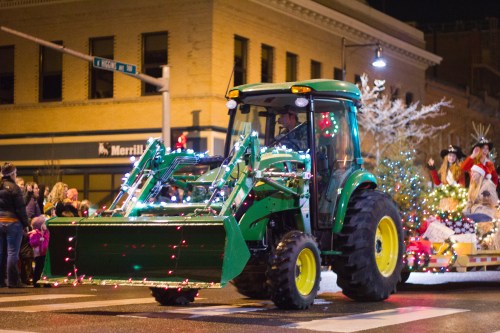 A decorated tractor with lights drives through a festive parade, surrounded by onlookers and holiday decorations.