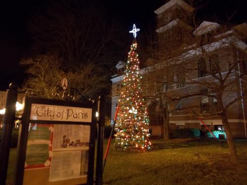 A decorated Christmas tree with a star stands beside a sign for the City of Paris, illuminated at night.