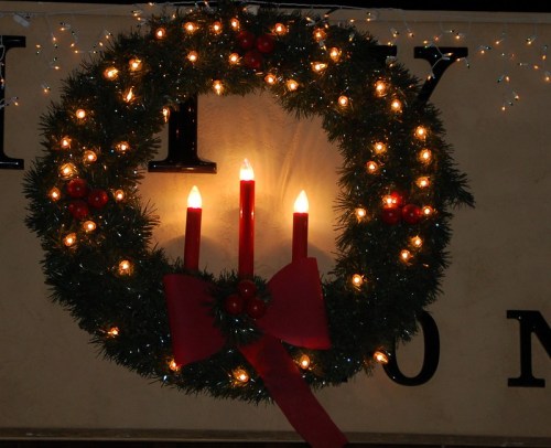 A festive wreath adorned with lights, red candles, and a bow, set against a wall with decorative lettering.