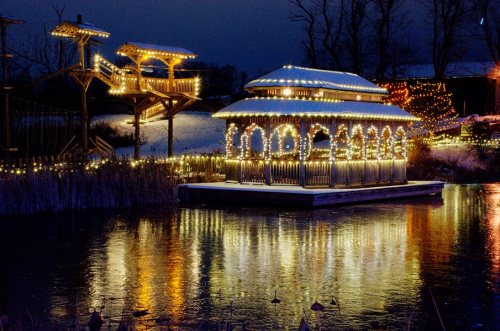 A beautifully lit gazebo reflects on a calm water surface, surrounded by trees and festive lights at dusk.