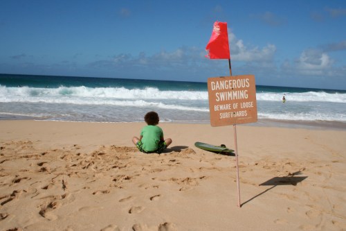 A child sits on the beach near a sign warning of dangerous swimming conditions and loose surfboards. Waves crash in the background.