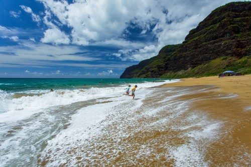 Two children play near the shoreline, splashing in the waves with a scenic beach and cliffs in the background.