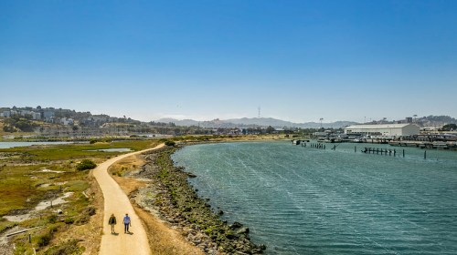 Two people walk along a scenic path by the water, with hills and buildings in the background under a clear blue sky.