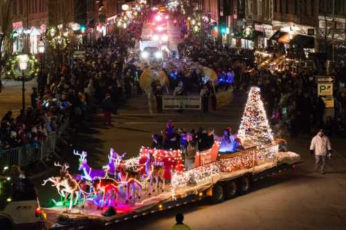 A festive parade scene with a decorated float featuring reindeer and a Christmas tree, surrounded by a large crowd.