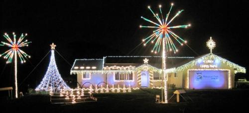 A brightly lit house decorated for Christmas with a large tree, starbursts, and colorful lights against a dark sky.