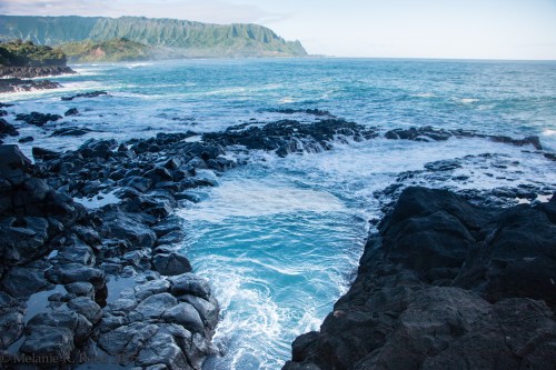 A rocky coastline with turquoise waves crashing against dark rocks, set against a backdrop of lush green mountains.