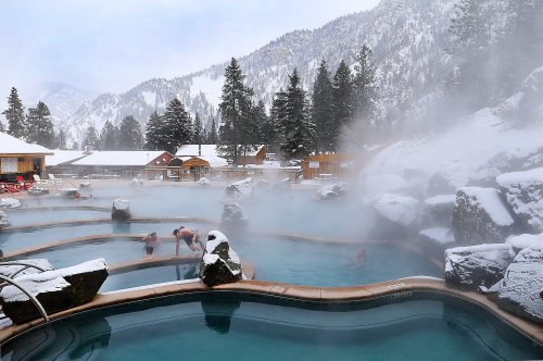 A scenic hot spring surrounded by snowy mountains and evergreen trees, with people enjoying the warm water.