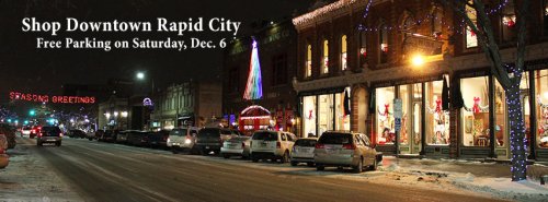 A festive downtown street in Rapid City at night, decorated with lights and holiday displays, with cars parked along the road.