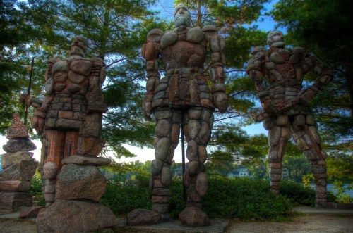 Three large stone statues resembling armored figures stand among trees, with a serene landscape in the background.
