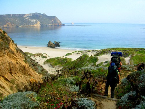 A hiker with a backpack descends a trail towards a serene beach, surrounded by lush greenery and rocky cliffs.