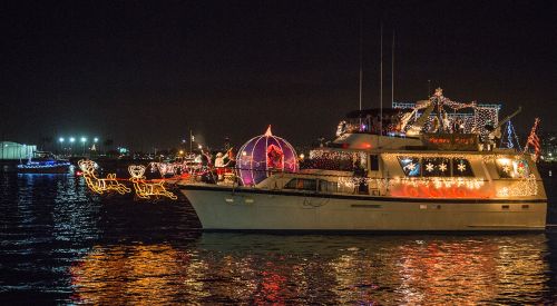 A decorated yacht with festive lights sails on a dark waterway, surrounded by colorful holiday displays.