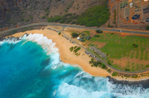 Aerial view of a beach with waves, parking area, and green spaces along the coastline.
