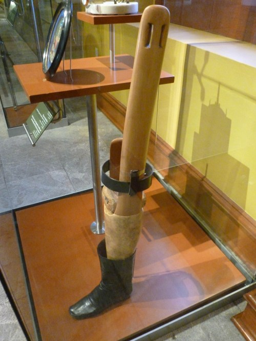 A wooden prosthetic leg displayed in a glass case, featuring a black leather boot and a strap for support.
