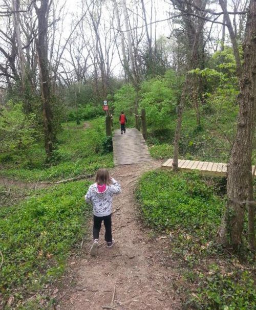 Two children walk along a wooded path, approaching a small wooden bridge surrounded by lush greenery.