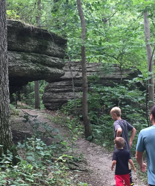 Two children walk along a wooded path with large rock formations and lush greenery surrounding them.