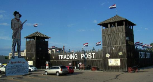 A wooden trading post with towers, flags, and a statue of a soldier in front, set against a clear blue sky.