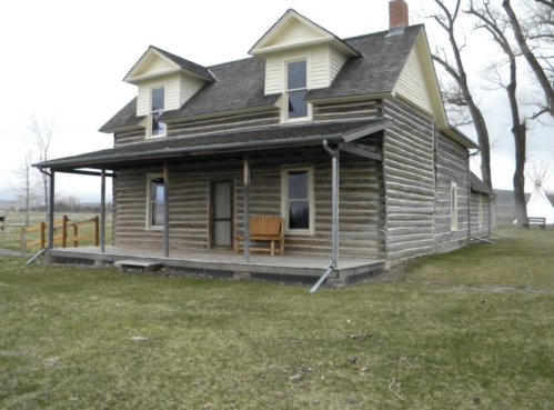 A rustic wooden house with a porch and two upper windows, set in a grassy area with trees in the background.