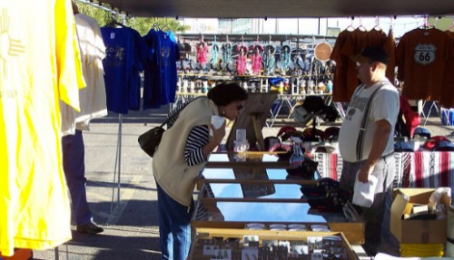 A woman examines items at a market stall, while a vendor stands nearby surrounded by clothing and merchandise.