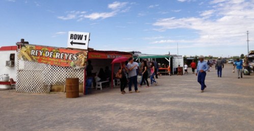 A bustling outdoor market with food stalls, people walking, and a sign pointing to "Row 1" under a blue sky.