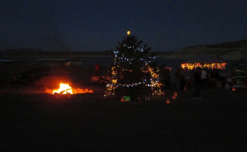 A decorated Christmas tree with lights stands near a bonfire, surrounded by festive decorations at night.