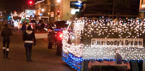 A festive parade scene with decorated vehicles and people walking, illuminated by colorful holiday lights.