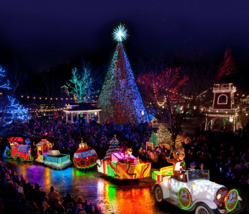 A festive parade with colorful floats and a large illuminated Christmas tree, surrounded by a crowd and twinkling lights.