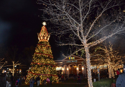 A brightly lit Christmas tree stands in a festive plaza, surrounded by twinkling lights and holiday decorations.