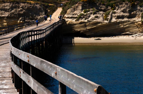 A winding wooden pier extends over calm water, leading to a sandy beach and rocky cliffs in the background.