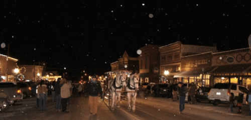 A snowy night scene of a bustling street with a horse-drawn carriage and people enjoying the festive atmosphere.